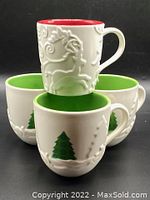 Four mugs arranged with one red interior embossed reindeer mug on top and three white mugs with green interiors and embossed Christmas tree scenes below.