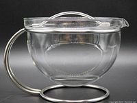 Gravy boat showing clear glass bowl resting inside round stainless steel base with attached handle and lid on top.