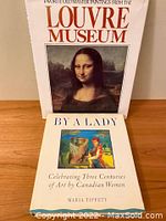 Two vintage art books standing side by side on wooden surface, showing front covers. One is Favorite Old Master Paintings from the Louvre with Mona Lisa on cover, the other is By a Lady: Celebrating Three Centuries of Art by Canadian Women with colorful female artist artwork on cover.