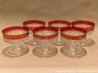 Set of six vintage Kings Crown Ruby Red low champagne glasses arranged in two rows on a surface highlighting their ornate red rim and clear thumbprint textured glass.