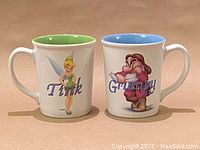 Front view of the two Disney character mugs, Tinkerbell and Grumpy, showing their character designs and names on white ceramic with colored interior.