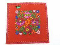 Front photo of red wool felt square with dense colorful Matyo style hand embroidery showing floral patterns and a signature foil leaf motif at top left corner.