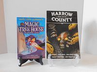 Two graphic novels standing on display stands: Magic Tree House and Harrow County Snake Doctor.