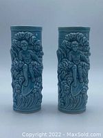 Front view of two blue tiki mugs showing surfing boys in 3-D relief against wave patterns