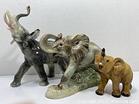 The three ceramic elephant figurines: large gray with raised trunk and pre-drilled holes, medium gray with iridescent finish and raised trunk, and small tan brown elephant with missing tusks, side view