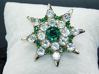Close-up image of the vintage rhinestone brooch showing the starburst design, green and clear rhinestones, and gold-toned metal prong settings.