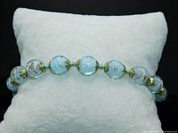 Full view of the bracelet displayed on white cushion showing round aquamarine Murano glass beads with gold foil swirl inside and gold flower bead caps, knotted with light blue thread between beads.