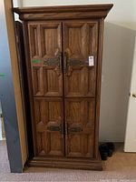 Front view of vintage solid wood armoire showing four paneled doors with ornate metal hardware and carved wood details.