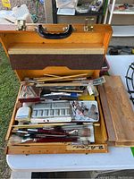 Wooden art supplies case open showing paintbrushes, chalk, paint palette, and paint tubes inside