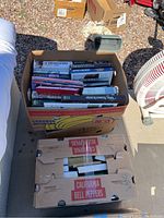 View of two cardboard boxes filled with assorted books including both hardback and paperback formats, photographed outdoors on concrete next to other items