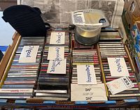 Large box filled with multiple rows of jewel case CDs with handwritten paper labels showing categories such as Opera, Broadway, Movies, Kids, Classics, and more. Blank CDs, JVC brand, and a black carrying case also seen in the box.
