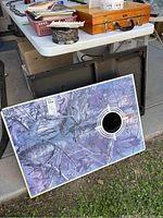 Two corn hole boards visible, one leaning against the other. The front board has purple-gray camouflage pattern and target hole.