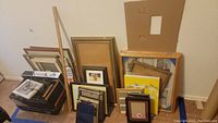 Wide shot showing a pile of assorted picture frames leaning against a wall, including large, medium, and small sizes with some framed art visible and many without glass.