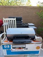 Silver and black HP Photosmart printer placed on original HP Home Photo Studio retail box outdoors, showing front and paper tray with some accessories.