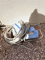 Blue EM 400 Hydra paint sprayer lying on gravel. Power cord coiled on top of unit, showing signs of wear and dirt.