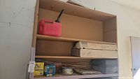 Wooden shelving unit with items including a red gas can, cardboard boxes, and some sandpaper or other flat materials stacked on shelves.
