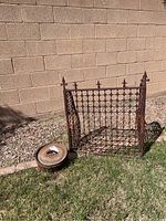 Side angled view of rusty brown cast iron grate with grid-like pattern and pointed finials, positioned on grass with a worn 10-pot beside it.