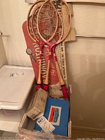 Three vintage wooden badminton rackets leaning on a wall with their original packaging, a Battleship game box, and a cardboard box with toy soldiers.