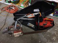 Two gasoline chainsaws, one inside a black hard plastic carrying case with manual, both placed on a concrete floor with surrounding tools and cords.