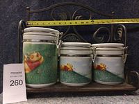 Photo of the set of three Pooh Bear ceramic cannisters of varying sizes secured on a metal shelf, with a tape measure showing approximate width of 12 inches.
