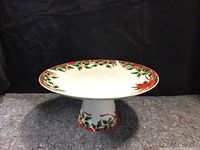 Front view of the white ceramic pedestal dish decorated with holly and red berries on the rim and base.