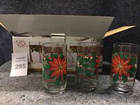 Photo shows three clear glass drinking glasses with red poinsettia and green leaf designs in front of an open box labeled 'Holiday Glasses' on a kitchen counter.