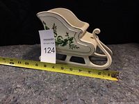Side view of white porcelain Christmas sled with green holly decoration and gold trim. Measuring tape shows length approximately 8 inches.