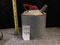 Front view of the vintage cylindrical metal fuel can showing ribbed body, red screw cap, angled spout, and measurement tape beside for scale.