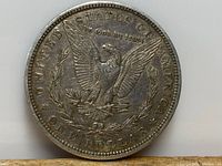 Reverse side of the 1887 American silver dollar coin showing the eagle with outstretched wings, 'In God We Trust', and 'One Dollar' inscriptions.
