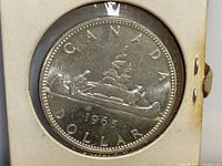 Obverse side of the 1965 Canadian silver dollar showing the voyageur and Indigenous person in a canoe with trees in the background.