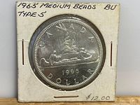 Obverse and reverse view of 1965 Canadian silver dollar coin with two people in a canoe, medium bead edge, in white holder with handwritten label.