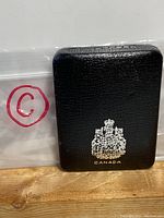 Black presentation case with embossed Canadian coat of arms and 'CANADA' text, placed on wooden surface, accompanied by plastic bag marked with 'C'.