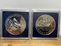 Front and back views of two Canadian silver dollar coins in protective cases. The left coin is Edmonton 1983, with a sport theme; the right is Detroit De Davis Strait 1987 with a sailing ship.