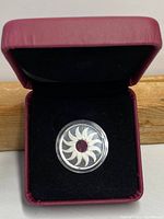 Silver coin with sun design featuring red gemstone center in protective clear case, displayed inside maroon and black velvet box.