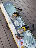 Burton Troop 116 snowboard lying on top of its soft carrying case with yellow and black mounted bindings, pair of red and white cracked goggles, and extra cleats arranged next to the board.