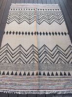 Full length view of the rectangular jute rug showing the black geometric diamond and zigzag patterns on beige base with multicolor woven bands at edges with fringes.