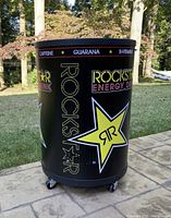 Side view of a cylindrical Rockstar Energy branded cooler, showing logo and design on black exterior with caster wheels.