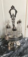 Front view of Victorian silver plate cruet set on pedestal stand showing four glass bottles with varied metal tops; one bottle missing stopper.