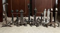 Full lineup of 11 various antique metal fireplace andirons showing a range of designs including twisted rods, flame motifs, geometric tops, and ball finials in black and white finishes