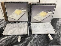Two boxed marble cheese cutting boards displayed side by side on a dark marble countertop. Left board missing wire, right board complete with wire and handle. Boxes shown behind.
