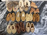 Photo showing full lot of 21 First Nations infant and child moccasins in various styles and materials, arranged on a marble surface.