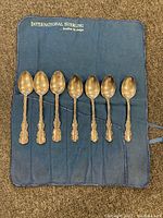 Seven sterling silver spoons arranged on a blue International Sterling cloth, showing the full set and ornate handle designs.