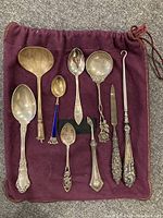 Top down photo of nine pieces of sterling silver flatware and serving utensils placed on a burgundy cloth pouch, showing all items with various decorative handle designs and styles.