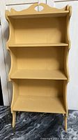 Front view of yellow painted wooden shelf showing three shelves and decorative top cut-out.