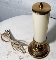 Unlit lamp showing ivory cylindrical glass shade, brass round base with embossed motif, tan power cord coiled beside base.