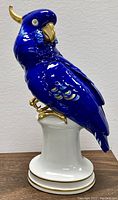 Front left side view of the blue and gold German porcelain parrot statue perched on a white cylindrical base with gold trim.