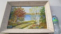 Framed oil landscape painting showing autumn trees, birch trees, a dirt path, and water in the distance.