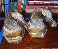Pair of metal horse head bookends displayed side by side on a wooden surface with books in the background.