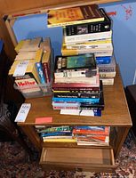 Books stacked on a wooden side table and below it, including paperbacks and hardcovers, showing some wear and yellowing on pages.