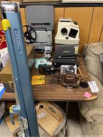 Wide view of lot showing all items on table including reel to reel recorder, cameras, projector, movie screen